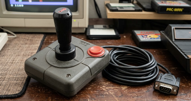 Joystick clásico de base cuadrada con palanca negra, botón rojo de disparo y la palabra FIRE en la carcasa, sobre una mesa de madera, con el cable enrollado y el conector DB9 macho bien visible a la derecha.